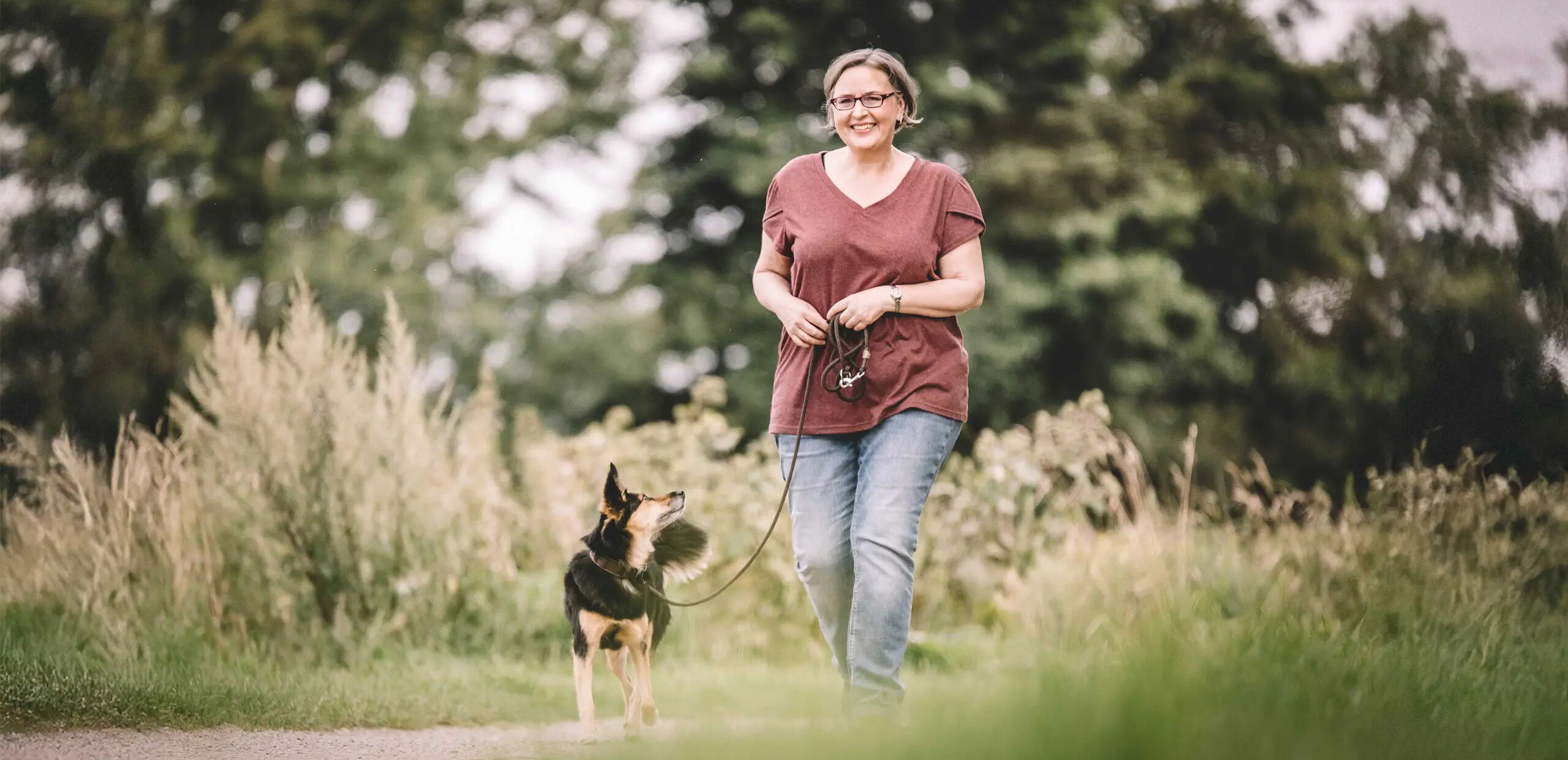 Eine Person geht mit einem Hund spazieren. Sie trägt ein rotes T-Shirt und blaue Jeans. Der Hund ist an der Leine. Der Hintergrund besteht aus grünen Bäumen und Gras. Die Szene wirkt ruhig und natürlich.