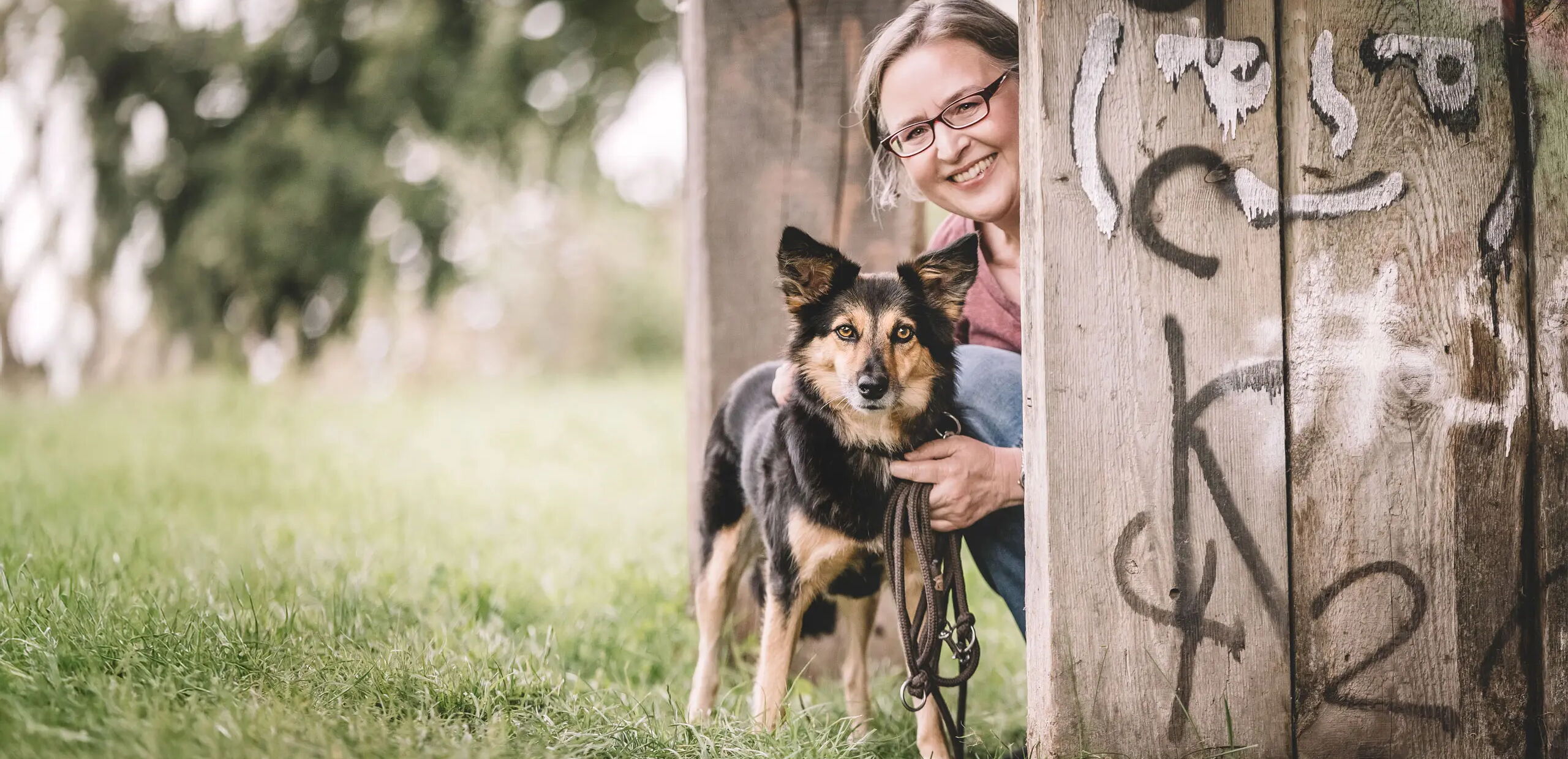 Eine Person kniet neben einem Hund unter einer Holzkonstruktion. Beide schauen in die Kamera. Die Holzbalken sind mit Graffiti bemalt. Im Hintergrund ist grüne Wiese und unscharfe Bäume zu sehen.
